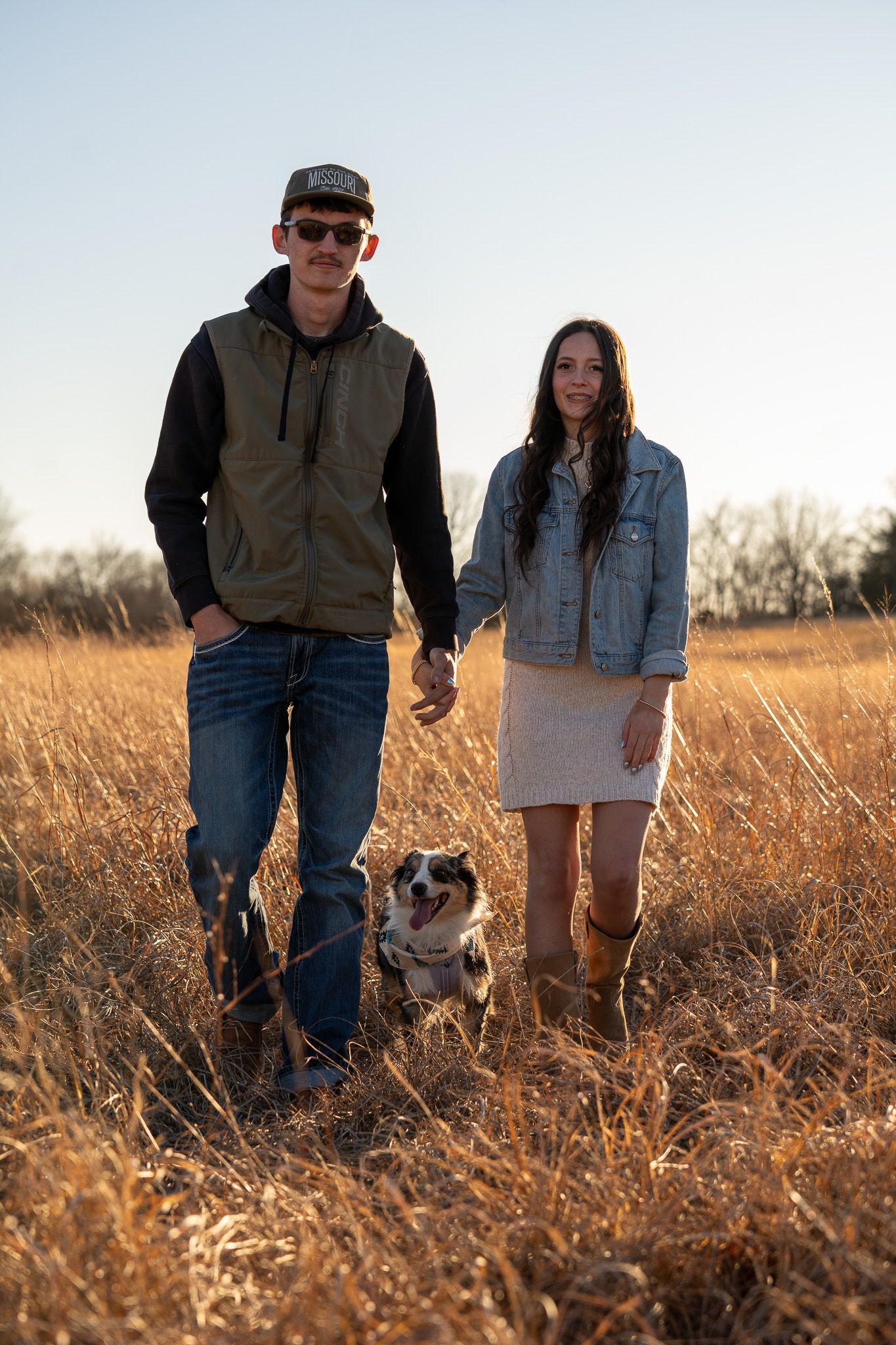 A couple walks through a golden wheat field at sunset with their dog, creating a warm autumn scene.