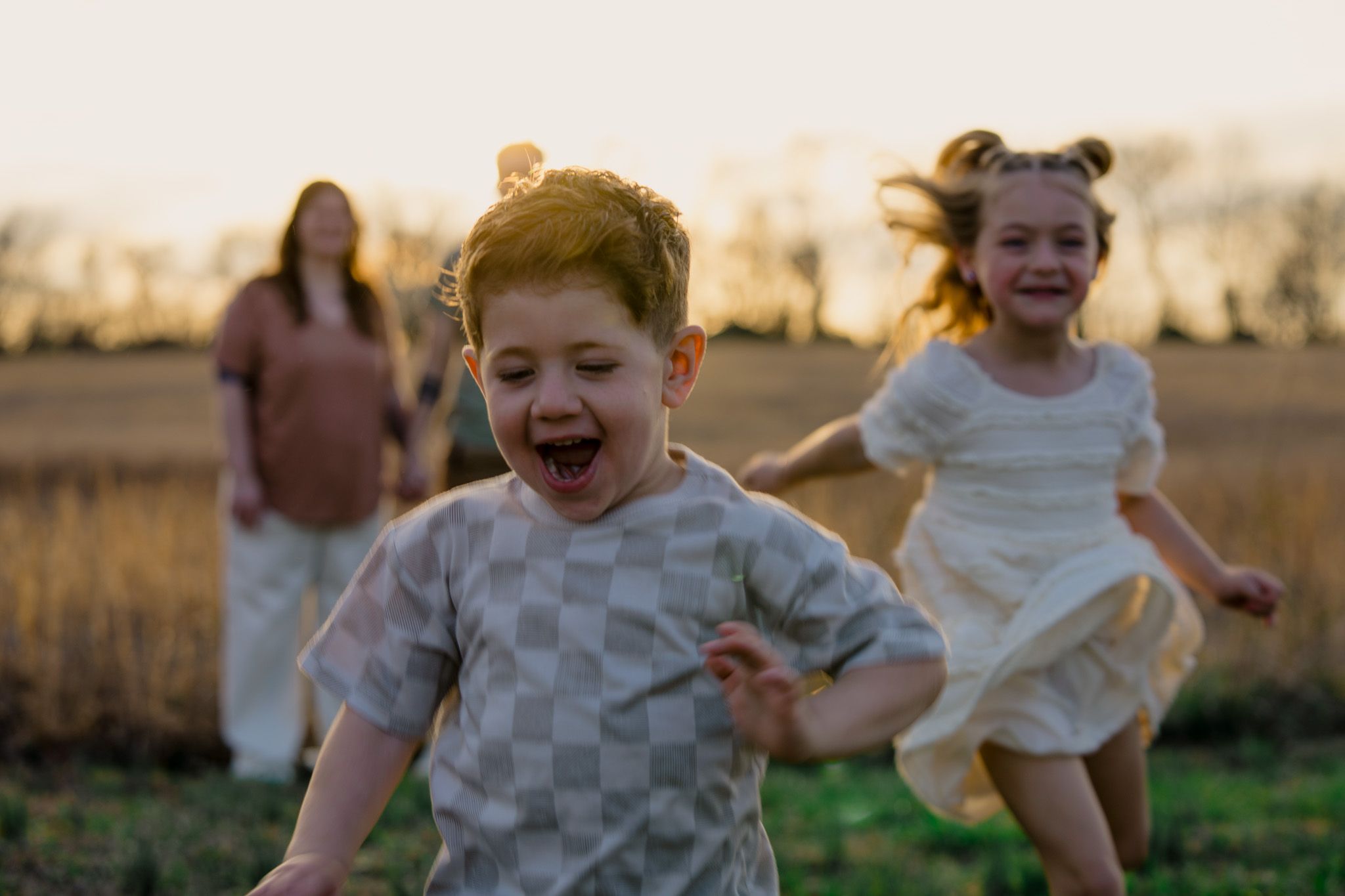 Children running and playing together in a golden field at sunset while an adult watches in the background.