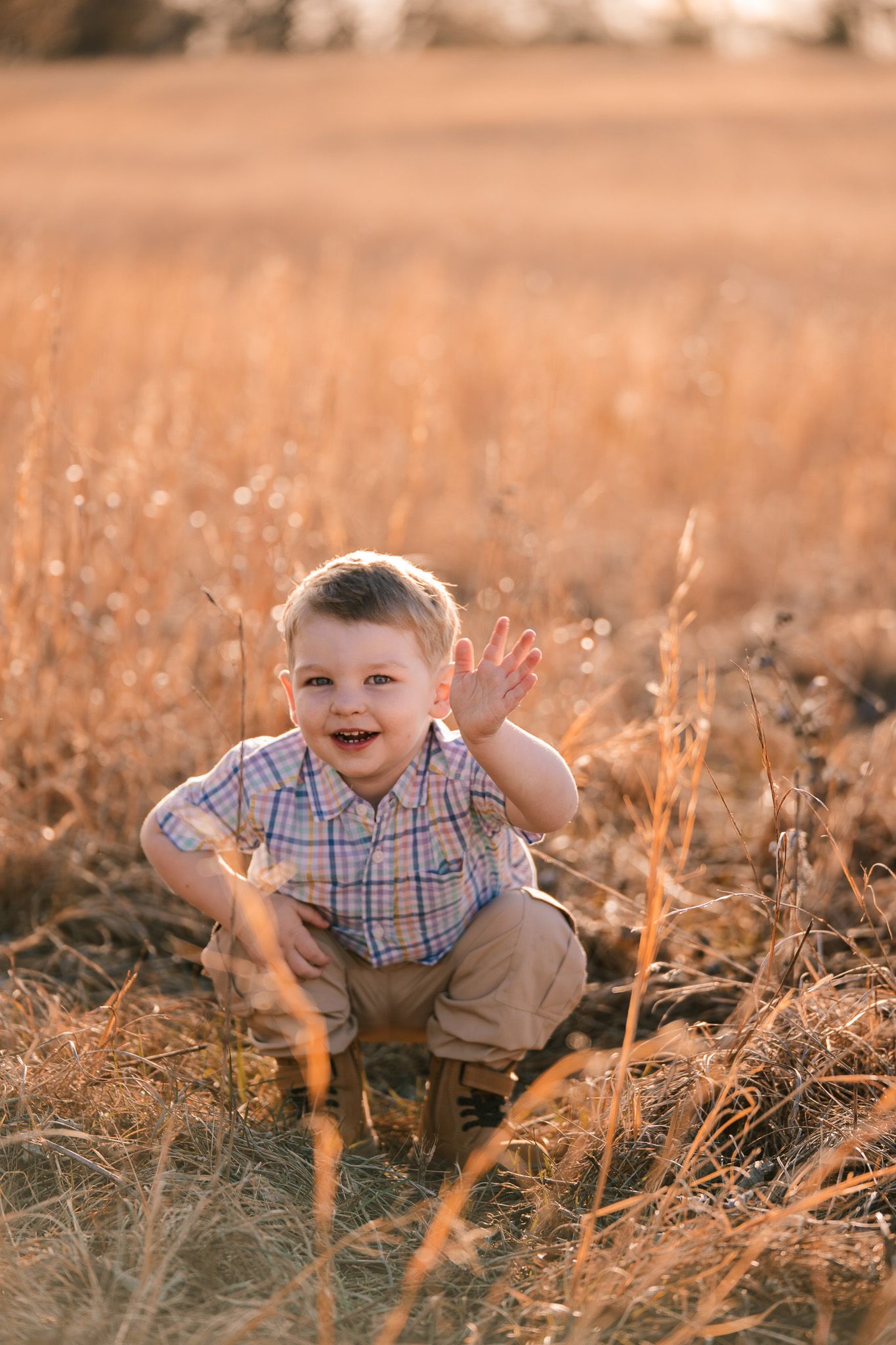 A young child plays and smiles in a golden wheat field at sunset, wearing a plaid shirt and khaki shorts.
