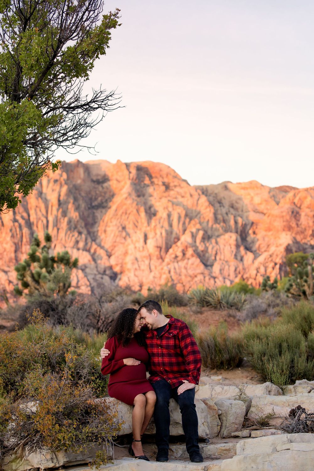 A couple sits together on desert rocks at sunset with red mountains glowing in the background.