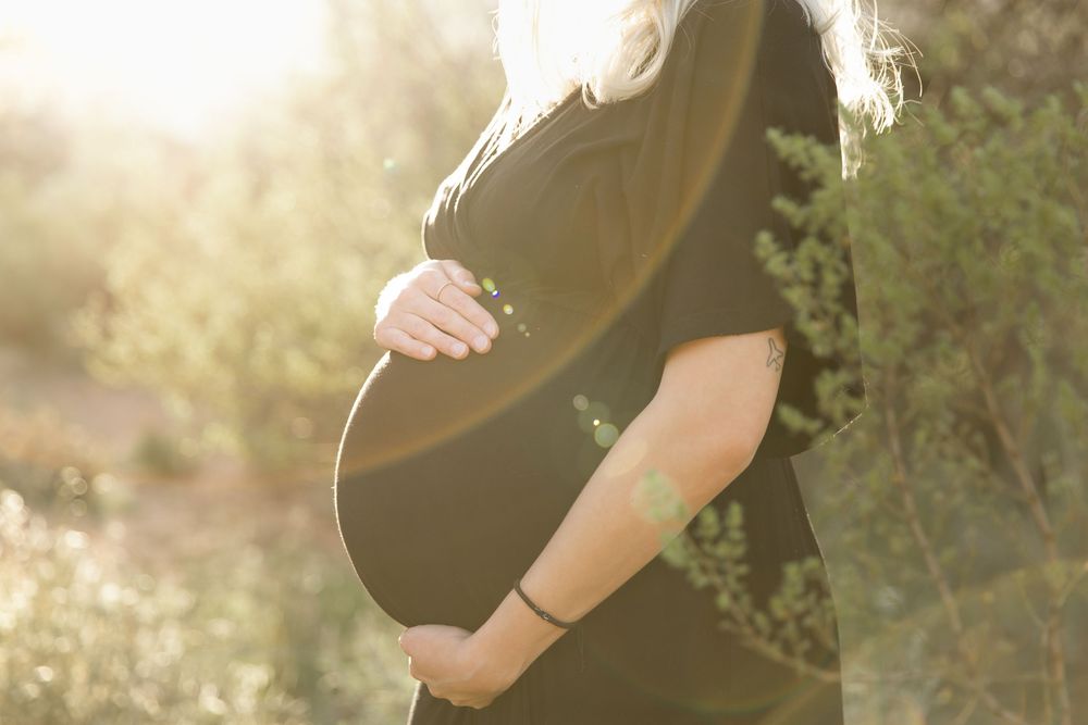 A pregnant woman in a flowy black dress poses in a sunlit outdoor setting during a maternity photoshoot.
