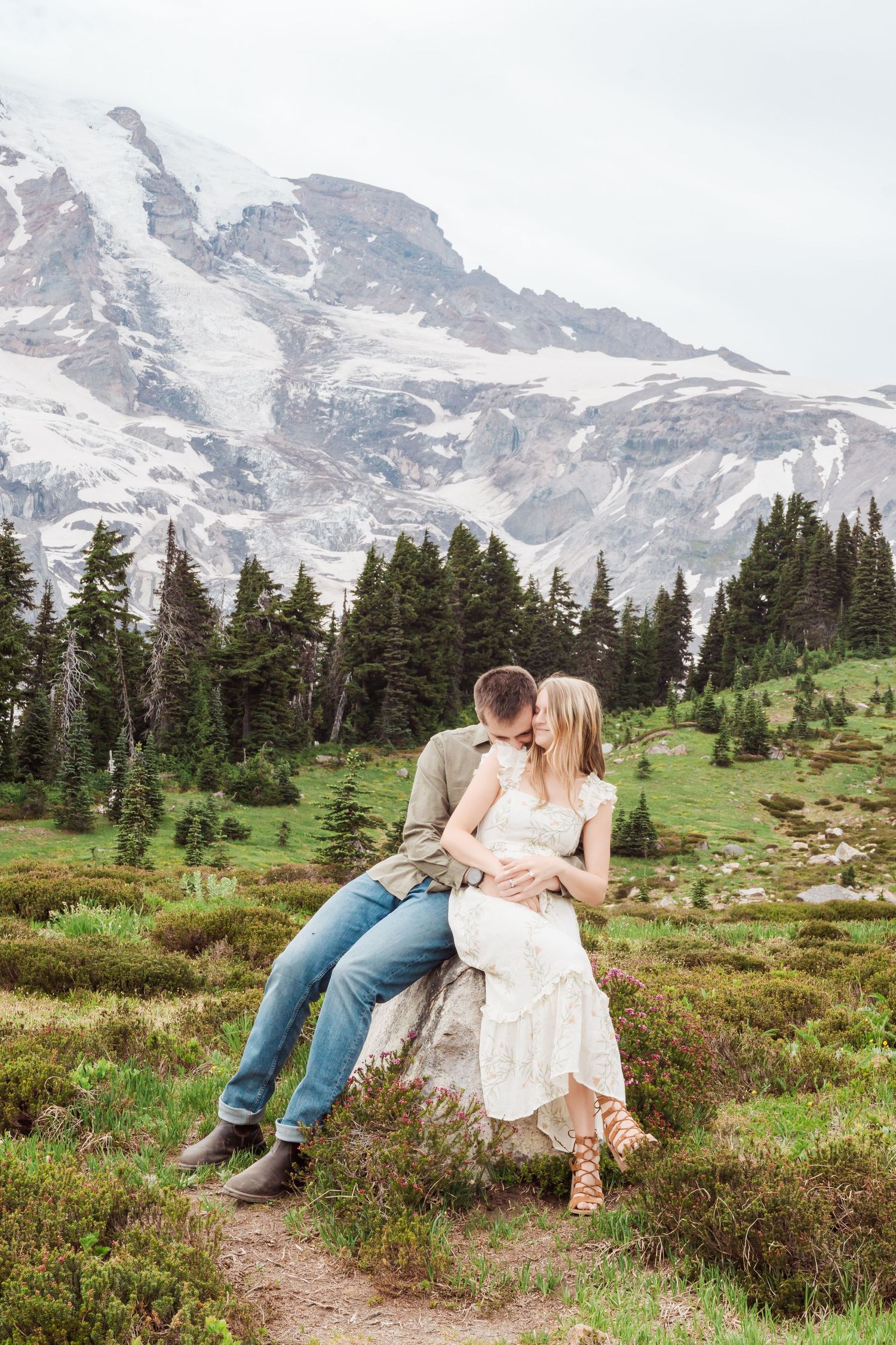 A couple sits on a rock in a meadow with Mount Rainier's snow-capped peak and evergreen forest in the background.