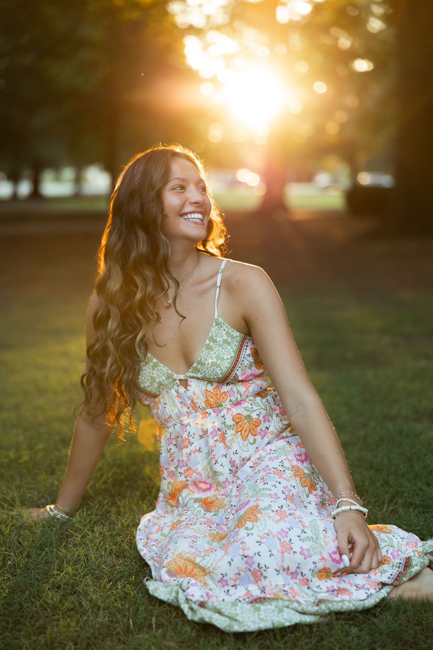 Woman in floral dress sits on grass during golden hour sunset.