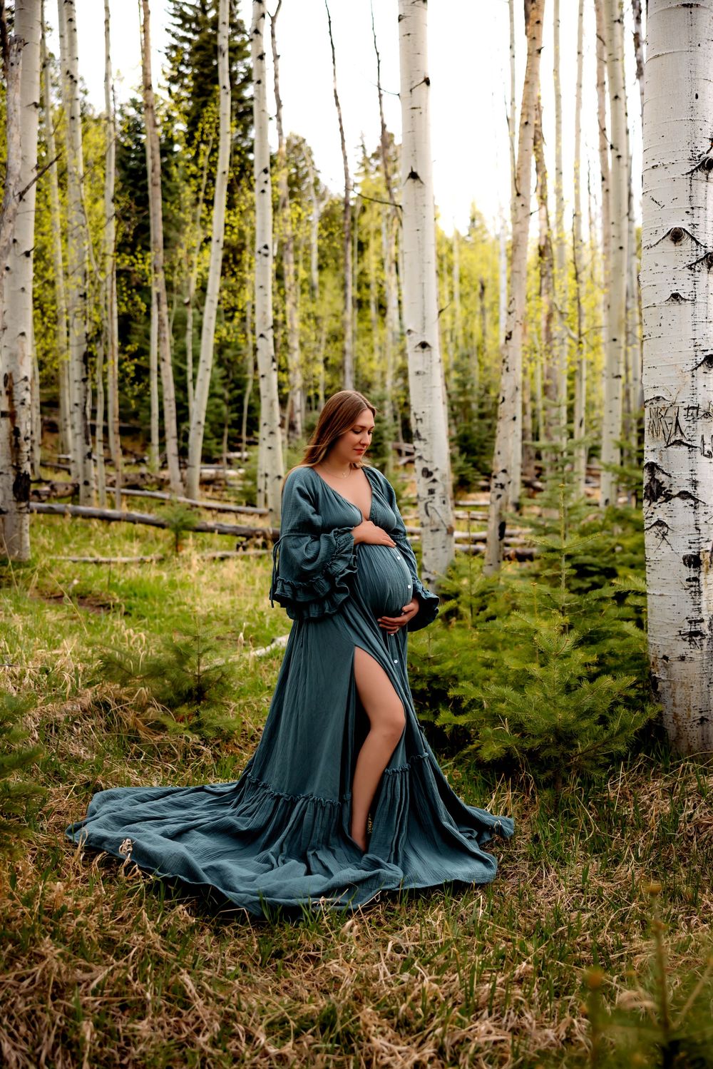 Pregnant woman in teal maternity gown stands among white birch trees in a lush forest setting.