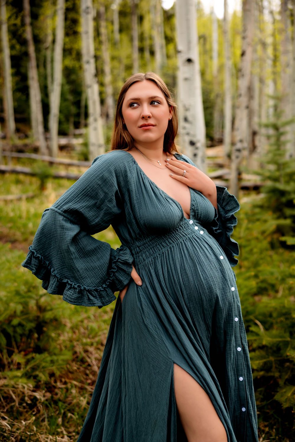 Pregnant woman in teal dress poses gracefully among aspen trees in a lush forest setting.