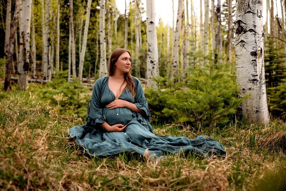 Pregnant woman in teal dress sitting peacefully among birch trees in a lush forest during a maternity photo session.