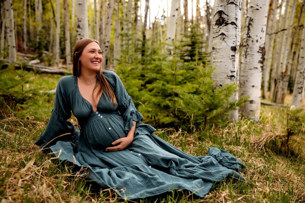 Pregnant woman in teal dress sitting among aspen trees in a forest, smiling and cradling her baby bump.