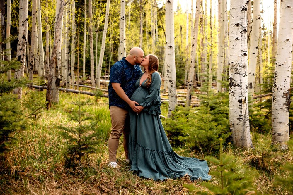 Couple sharing a tender kiss in a sunlit aspen forest, woman in flowing teal gown, maternity photo session.