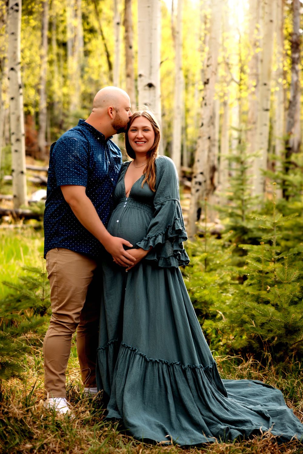 Couple poses in aspen forest, woman in teal maternity gown cradling baby bump while man kisses her cheek.