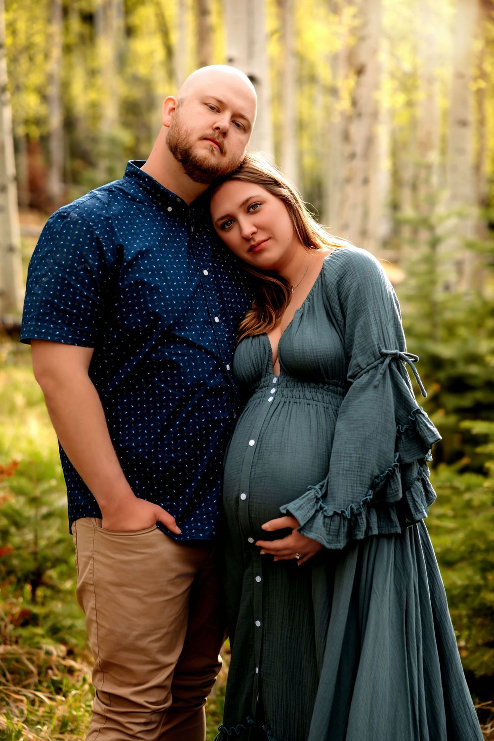 Expecting couple in teal and navy poses tenderly among golden aspen trees during maternity shoot.