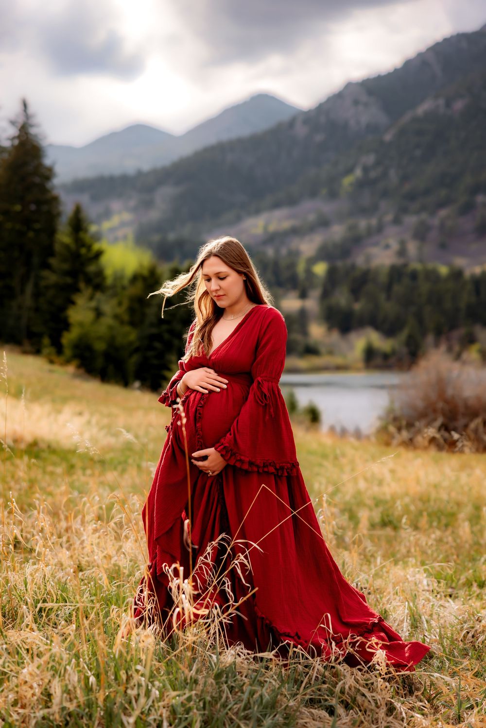 Pregnant woman in flowing red gown stands in mountain meadow with lake and peaks behind her.