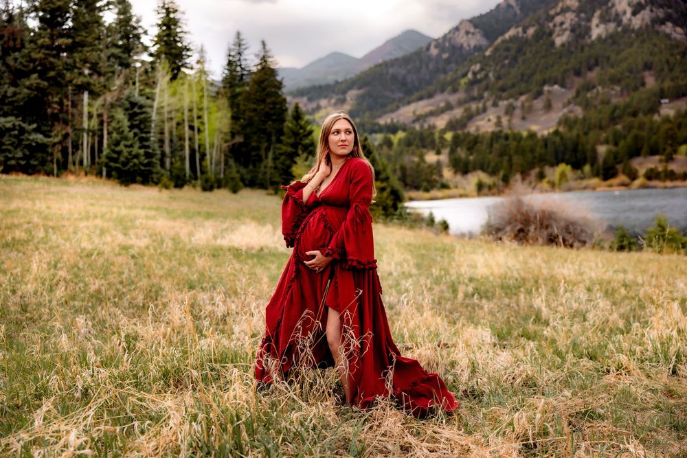 Pregnant woman in flowing red gown poses in mountain meadow with lake and pine trees backdrop.