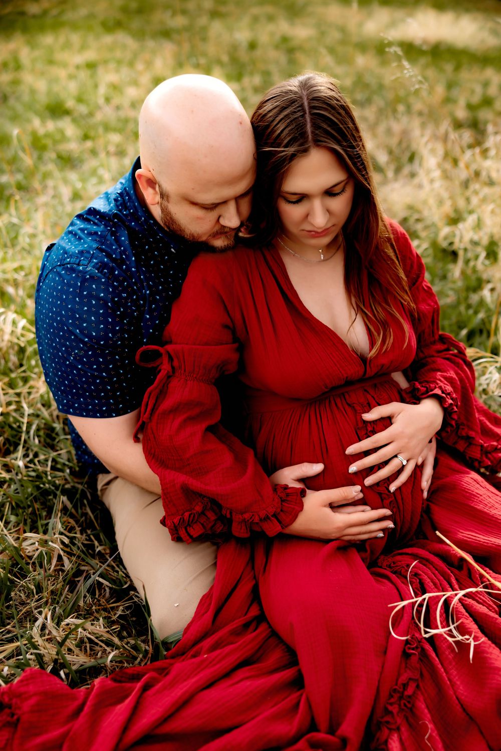 Expecting couple sitting in field, woman in flowing red maternity dress held by bald man in blue shirt, both gazing at baby bump.