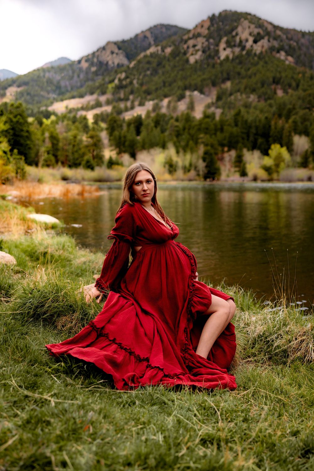 Pregnant woman in flowing red gown sitting by mountain lake with lush green hills backdrop.