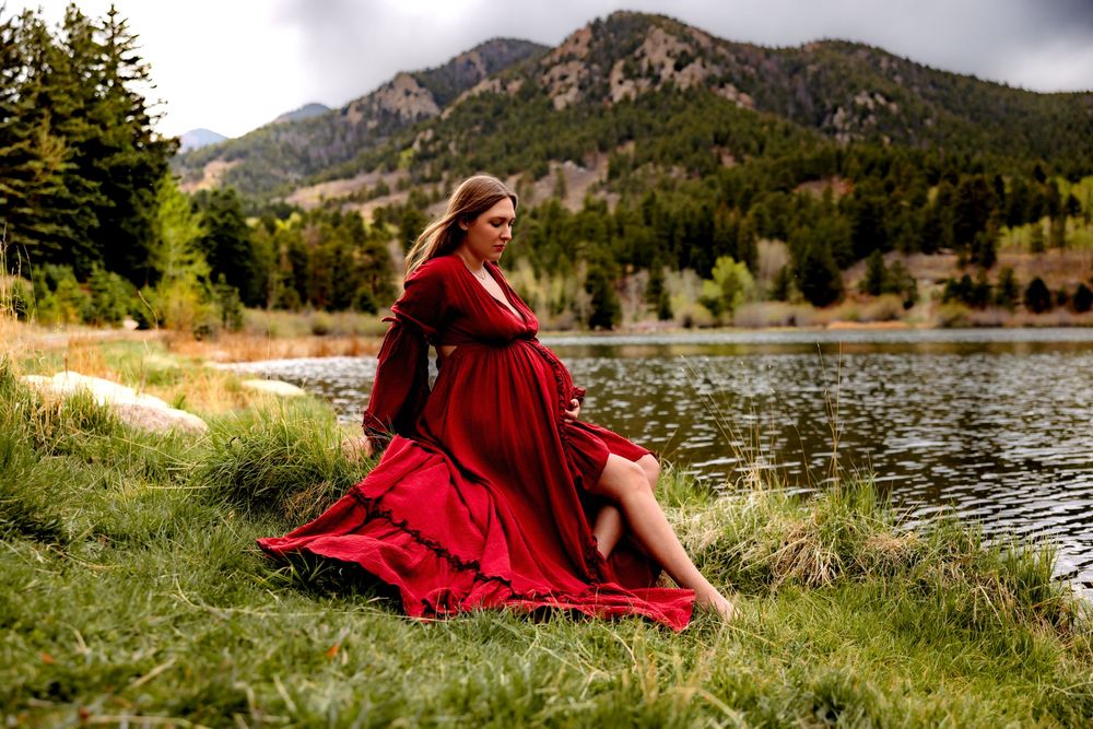 Pregnant woman in red dress sitting by mountain lake with evergreen trees and rocky peaks in background.