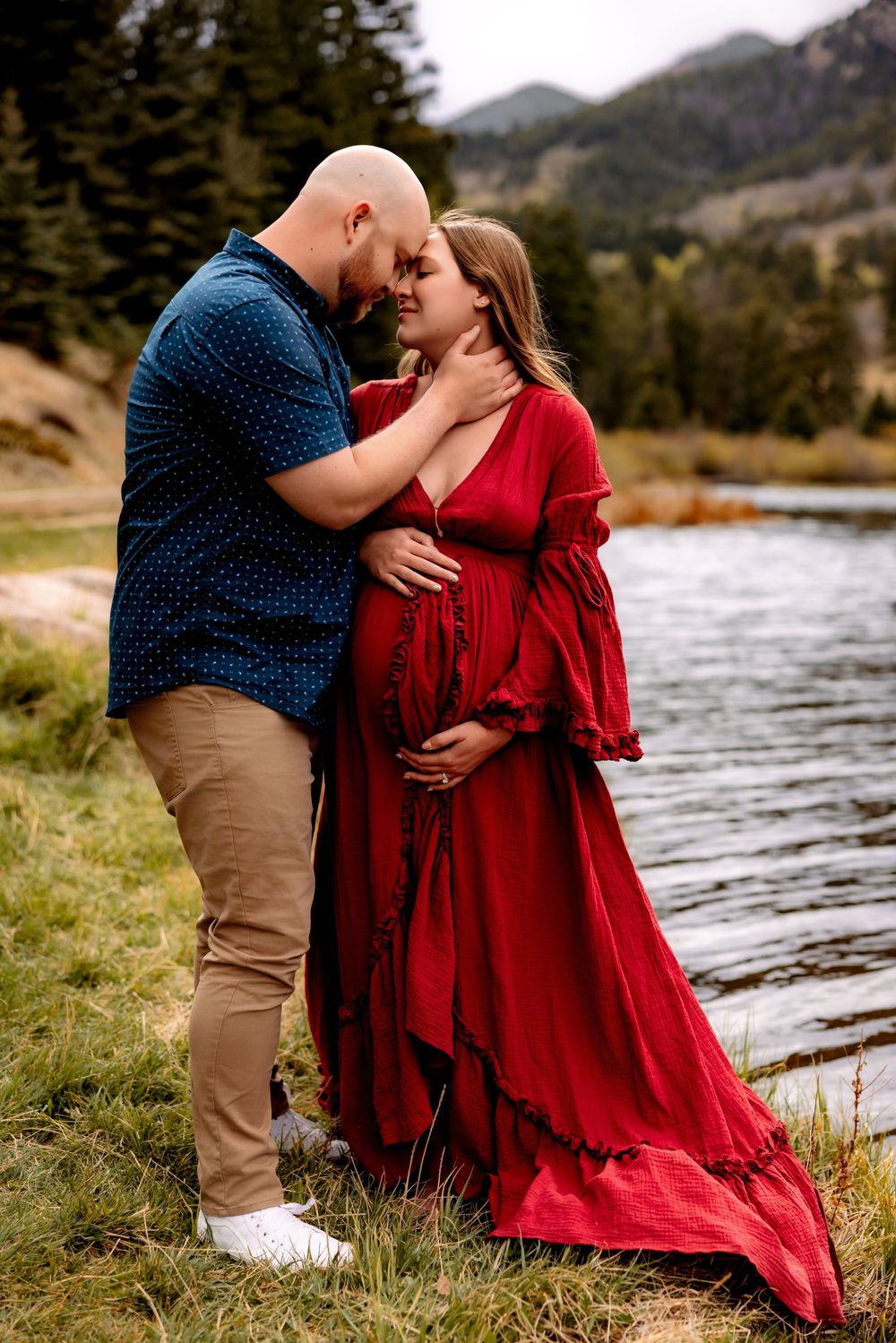 Couple sharing tender moment by mountain river, woman in red maternity gown, man in blue shirt.