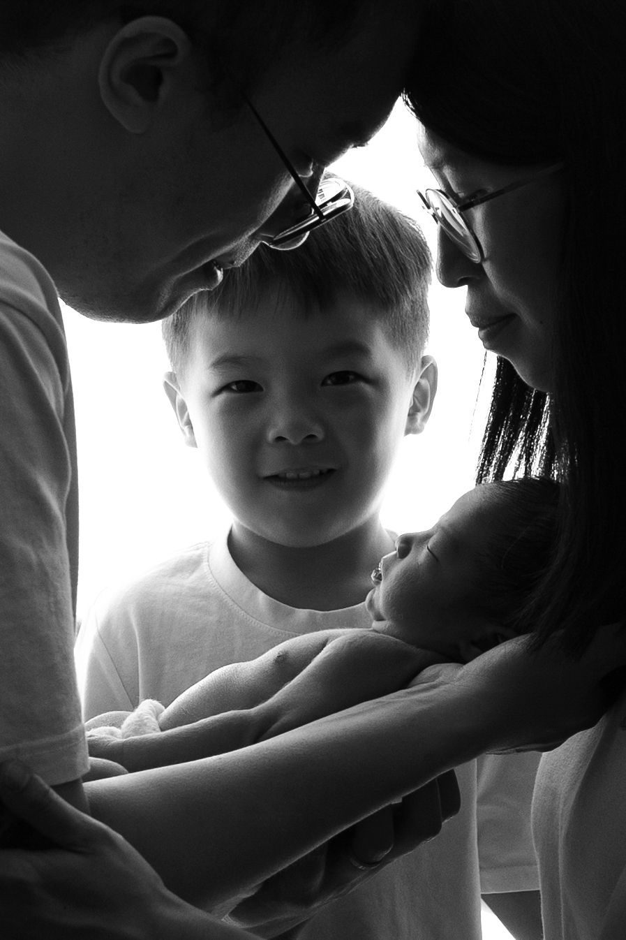 Black and white family portrait of parents with young boy and newborn baby, tender moment captured in soft light.