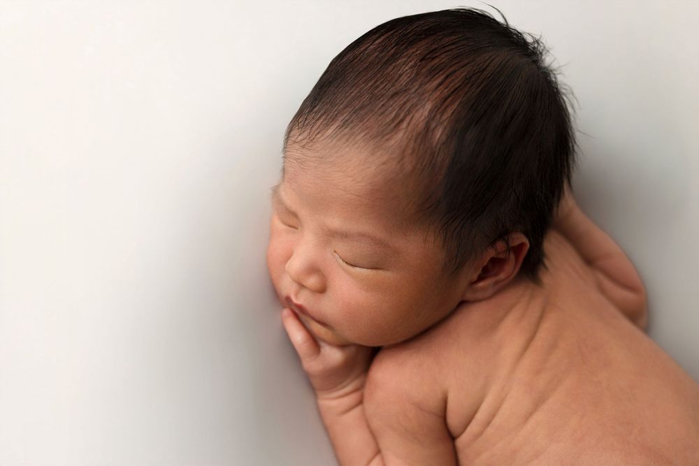 Peaceful newborn baby sleeping curled up with dark hair against a soft white background in a professional newborn photo.