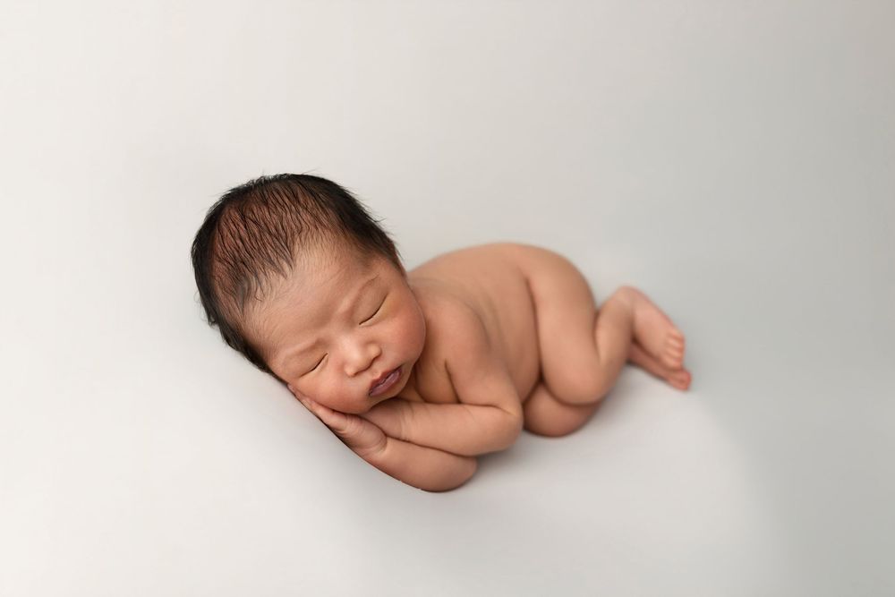 Peaceful newborn baby sleeping curled up on a soft white background in a classic posed newborn photography portrait.