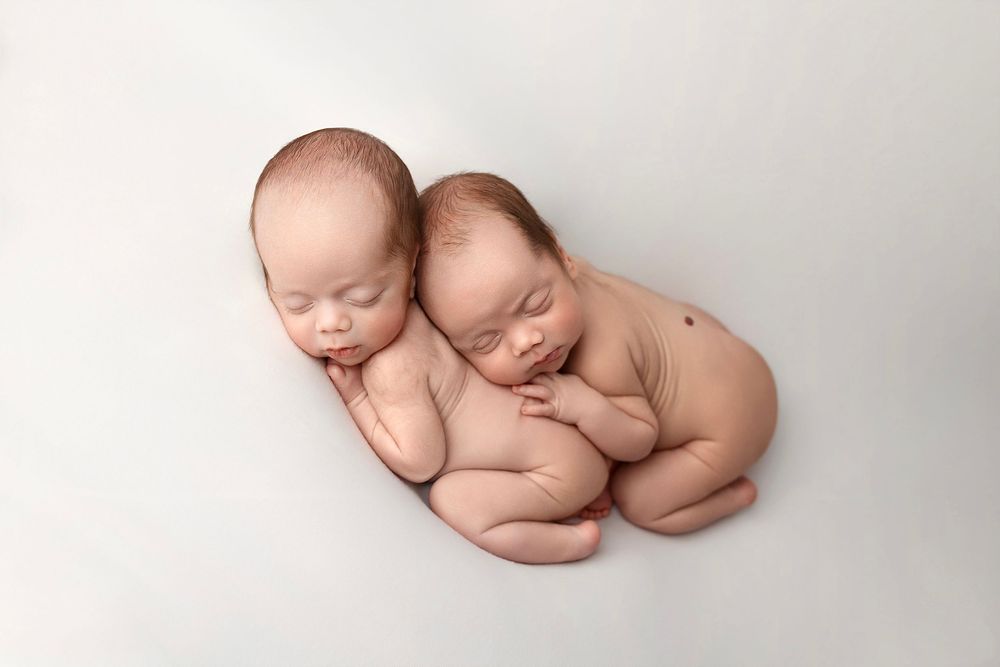 Two sleeping newborn babies curled up together on a white backdrop in a peaceful pose.
