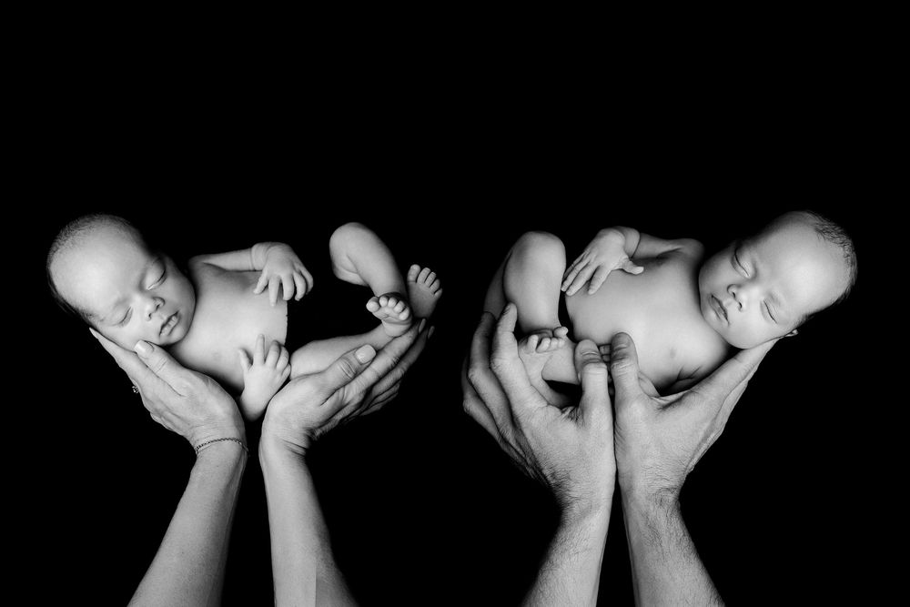 Black and white artistic portrait of two newborn babies being gently cradled in adult hands against dark background.
