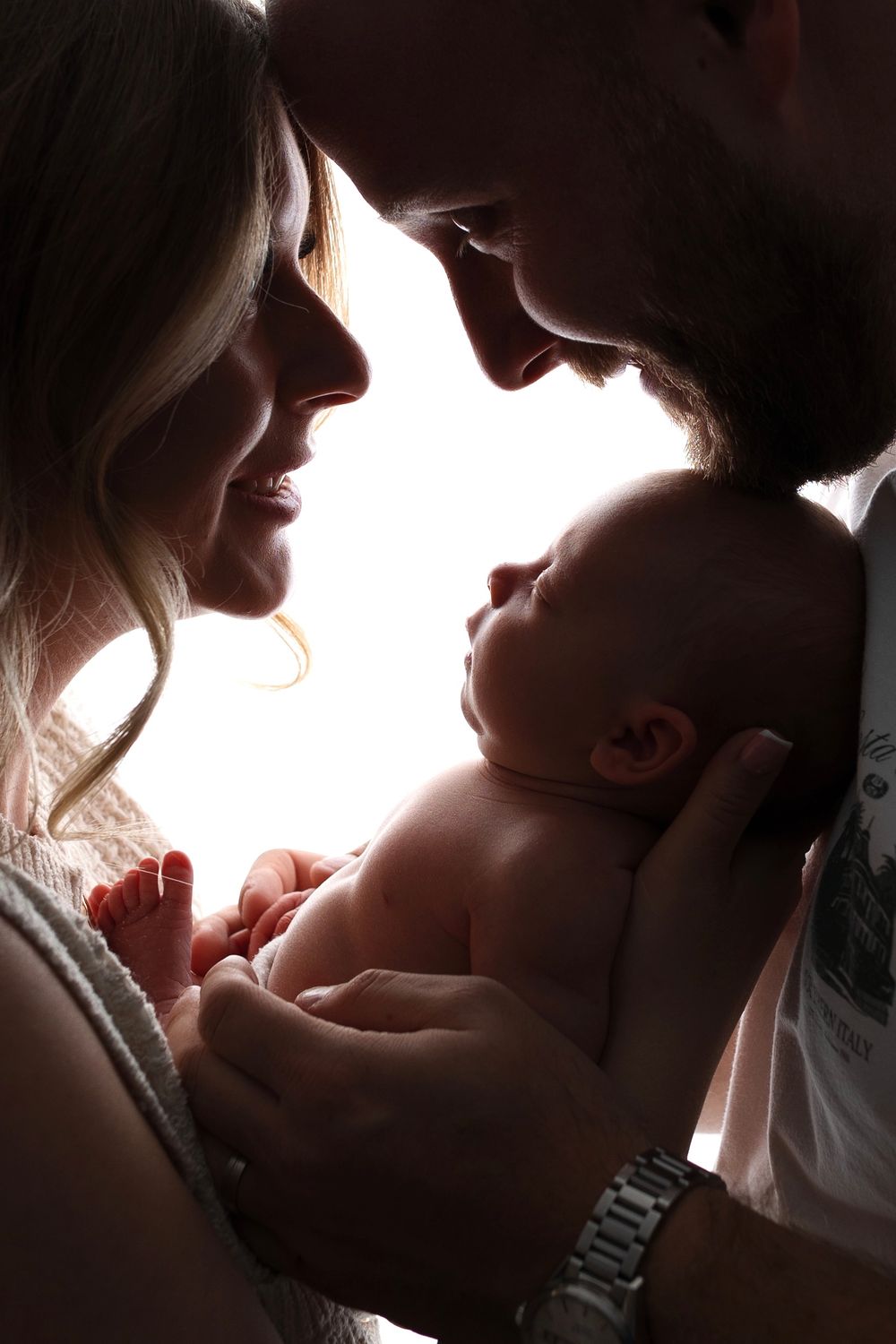Parents tenderly hold their newborn baby close, sharing an intimate family moment in soft backlit silhouette.