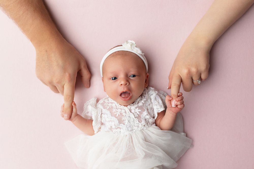 Newborn baby in white lace dress on pink background held by parents hands.