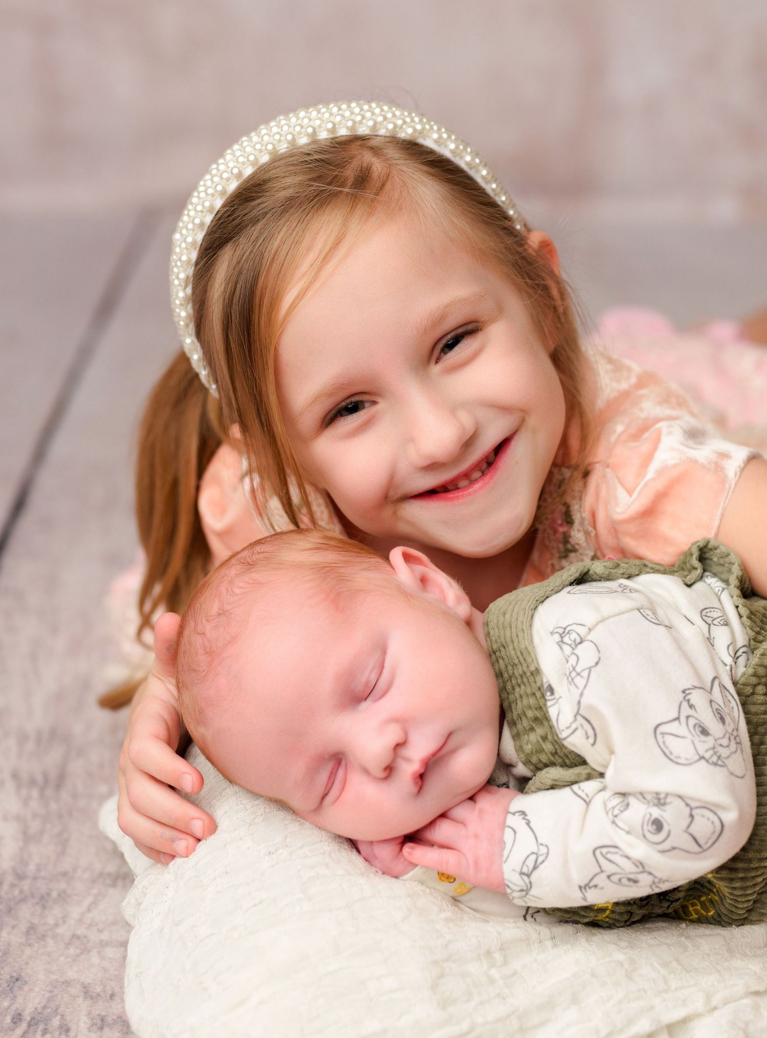 Smiling young girl with pearl headband cuddling sleeping newborn baby sibling in a sweet portrait photo.