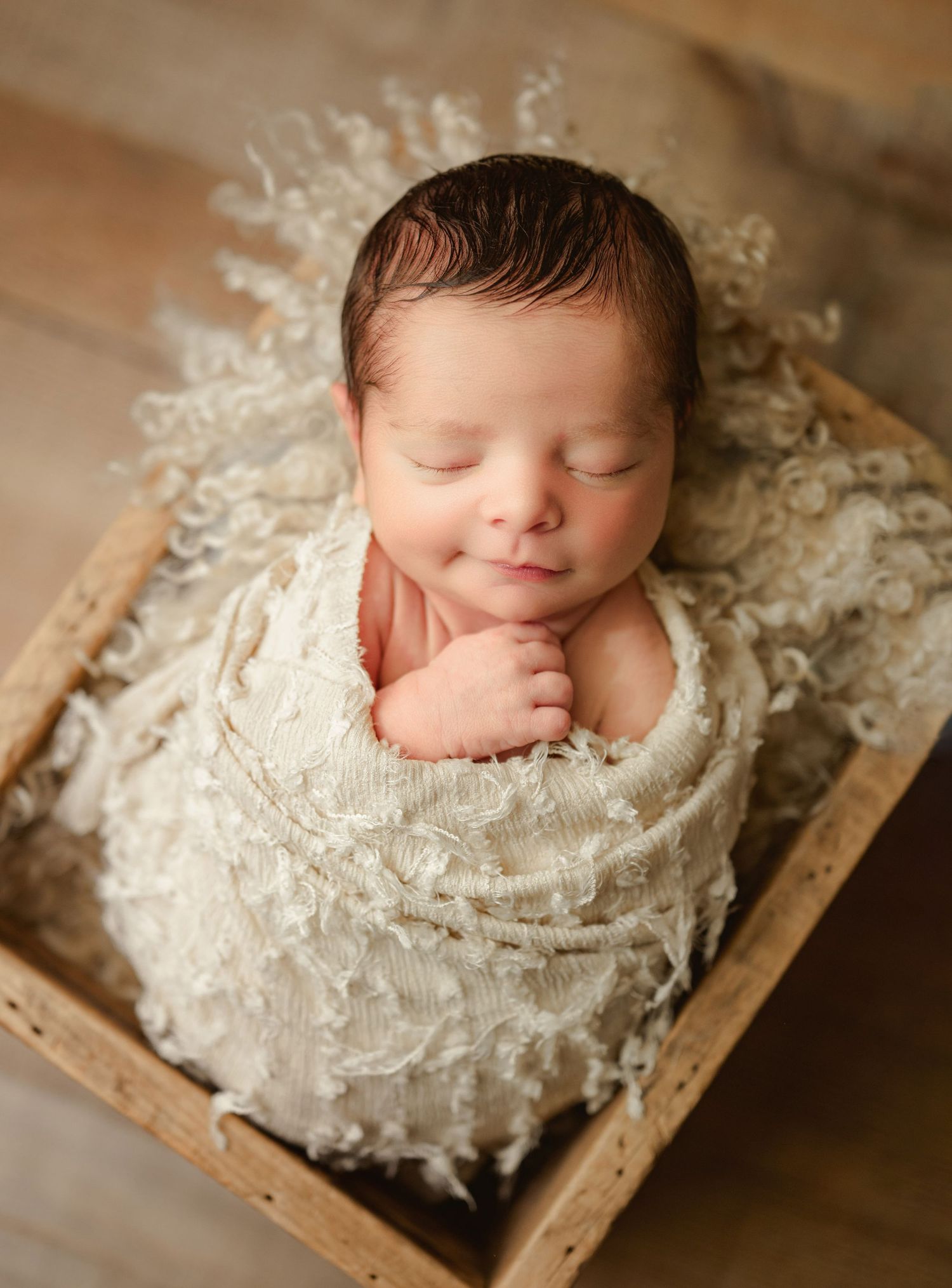 Sleeping newborn baby swaddled in cream wrap, nestled in rustic wooden box with fluffy white filler. Newborn Photography