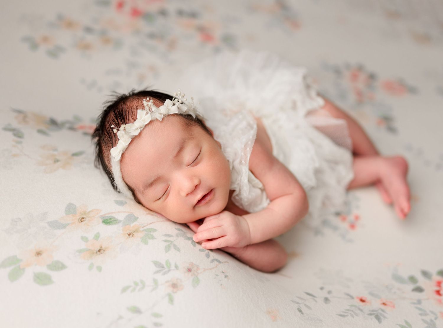 Sleeping newborn baby girl in white lace outfit and floral headband on a soft floral background. Newborn Photography in Buffalo