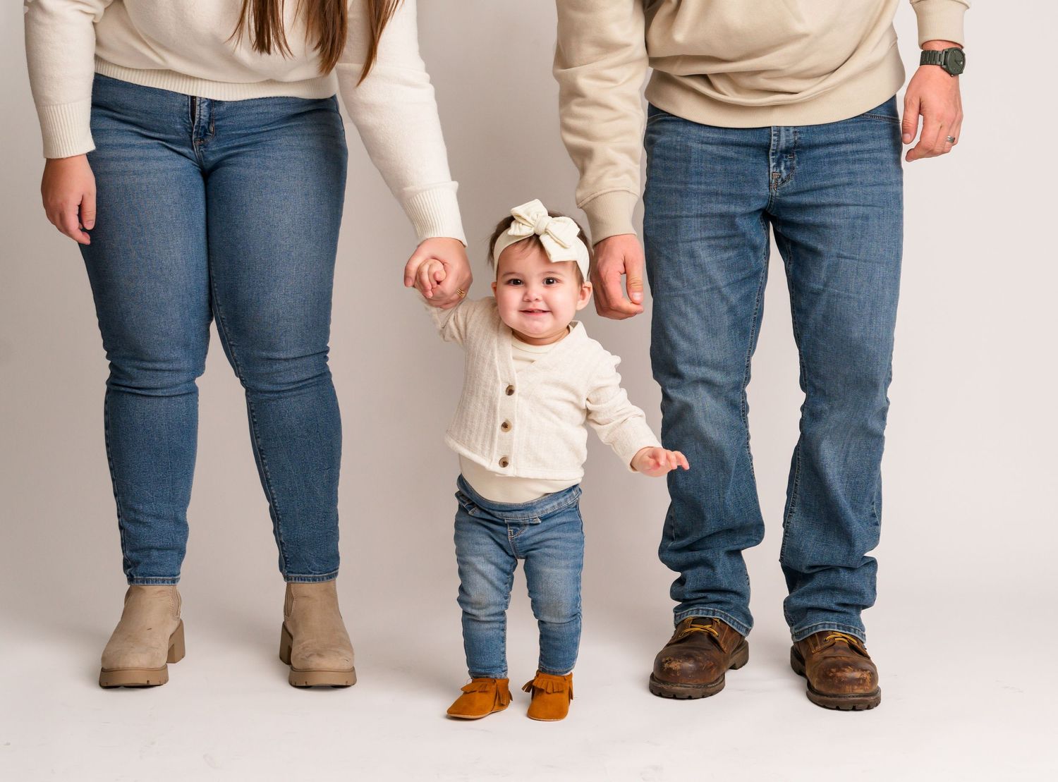 Smiling baby in white cardigan and jeans stands between parents holding hands in a studio photo. First Birthday Photography
