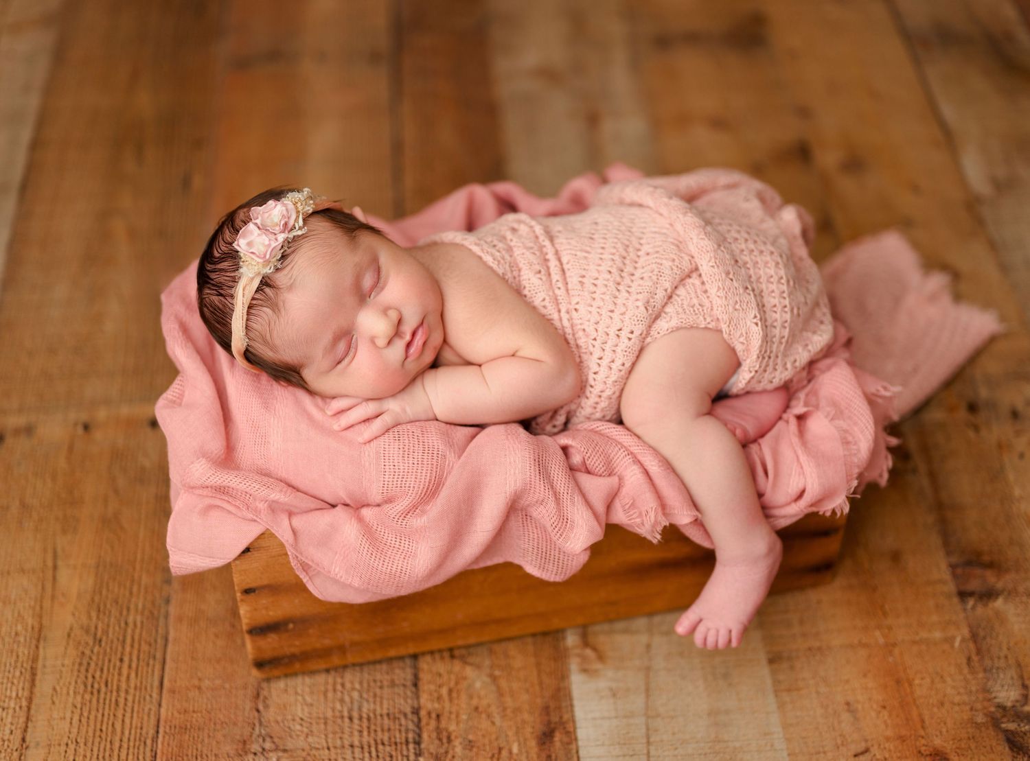 Newborn baby girl sleeping peacefully in a wooden box with pink knit wrap and floral headband on rustic wood floor.