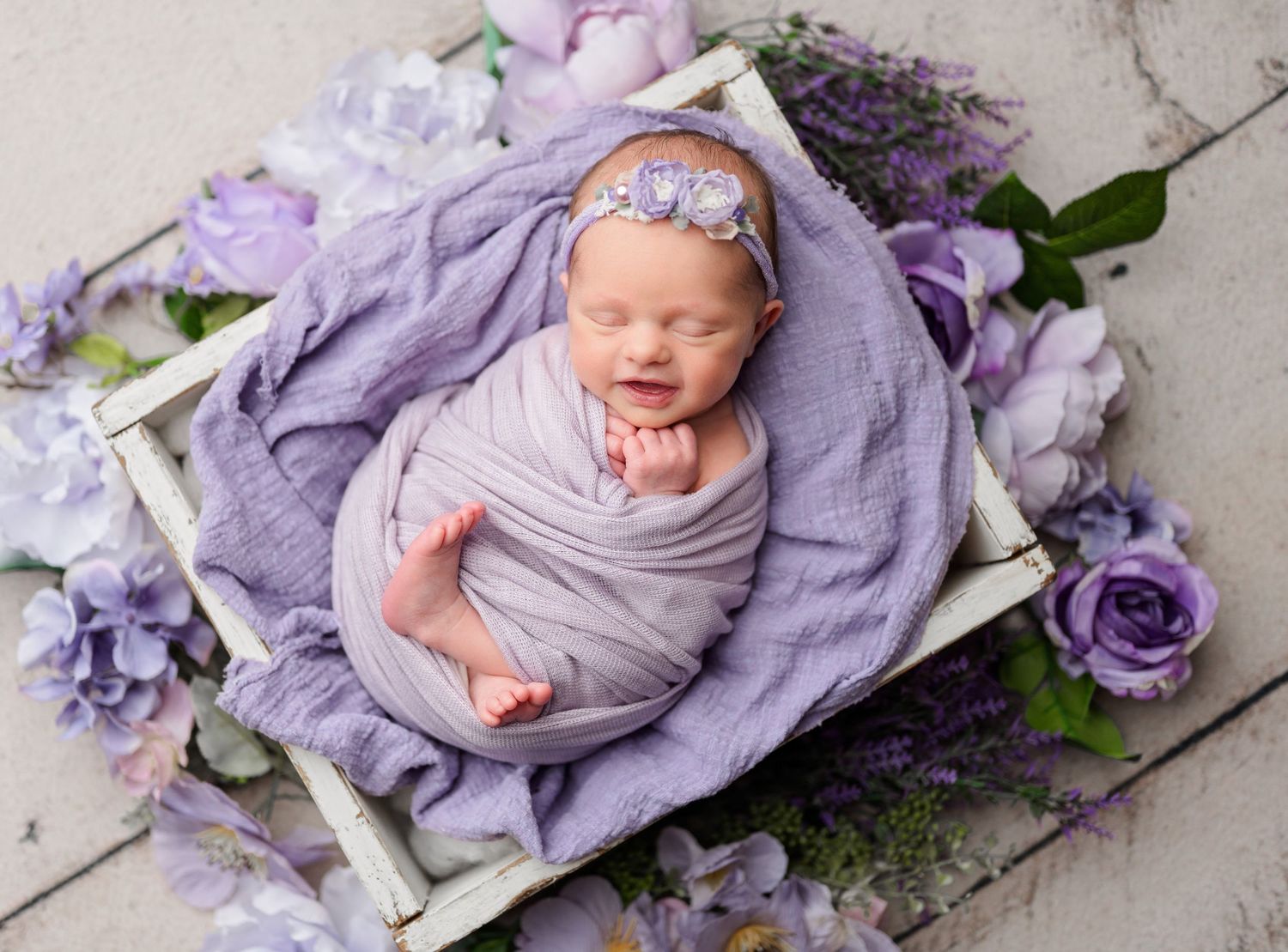 Smiling newborn baby wrapped in purple swaddle sleeping in white wooden crate surrounded by purple flowers. Newborn Photography in buffalo