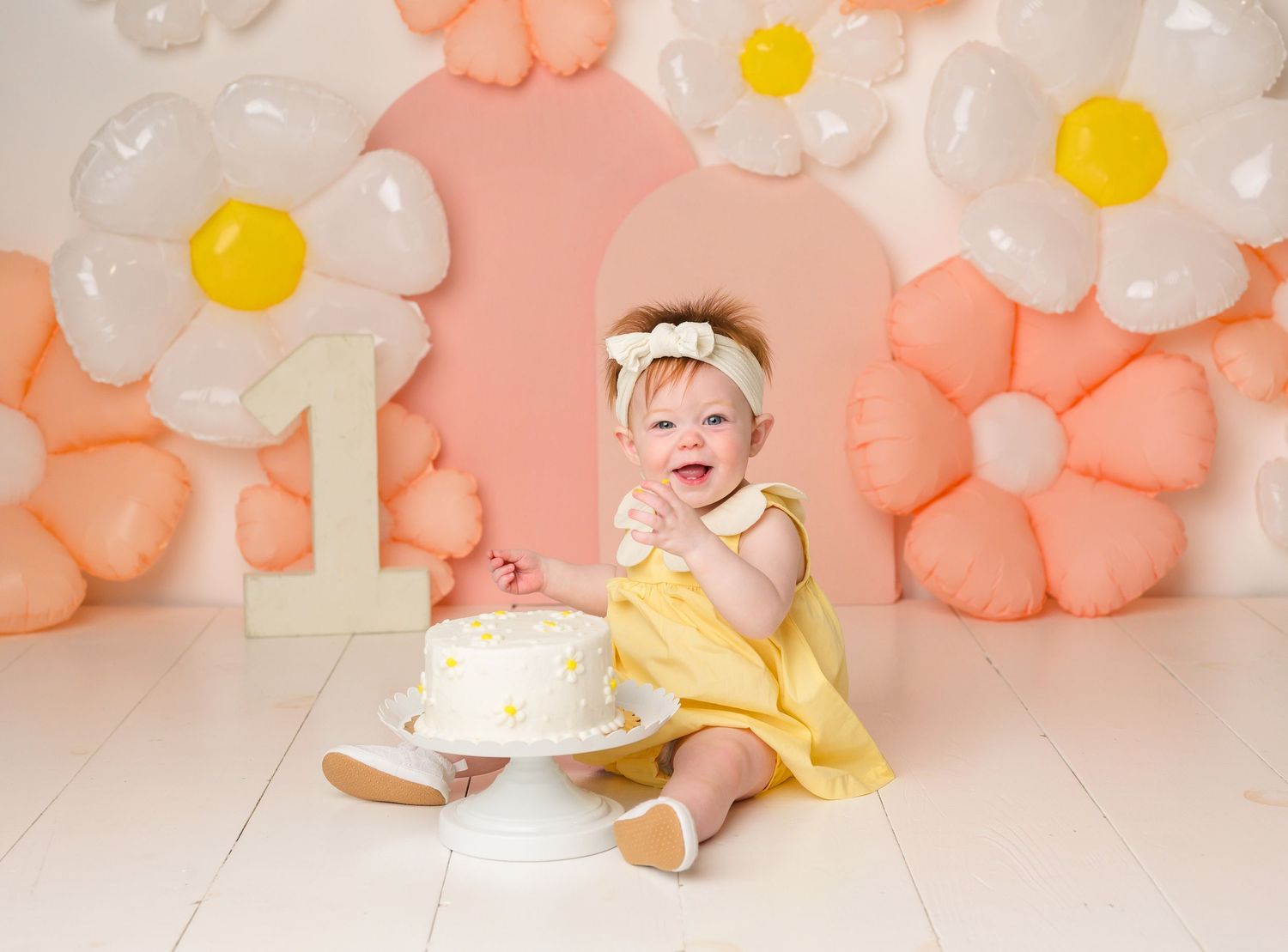 Baby girl smiles at first birthday cake smash with daisy balloon backdrop and number one decoration. First Birthday Photography