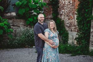 A maternity photo taken in front of a rustic brick wall with green foliage.