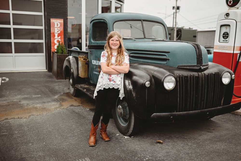 Young person poses confidently in front of vintage blue pickup truck at gas station.