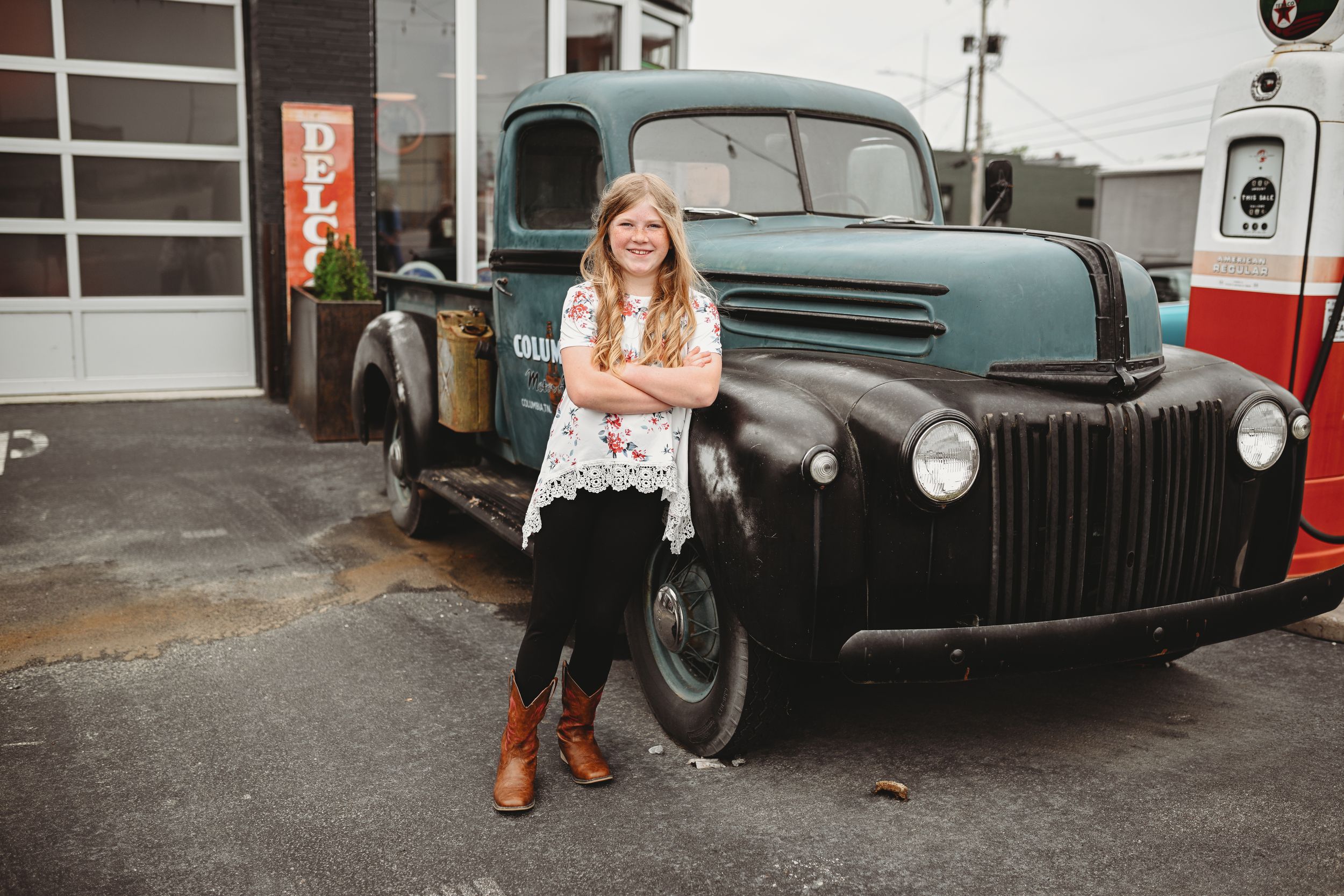 Young person poses confidently in front of vintage blue pickup truck at gas station.