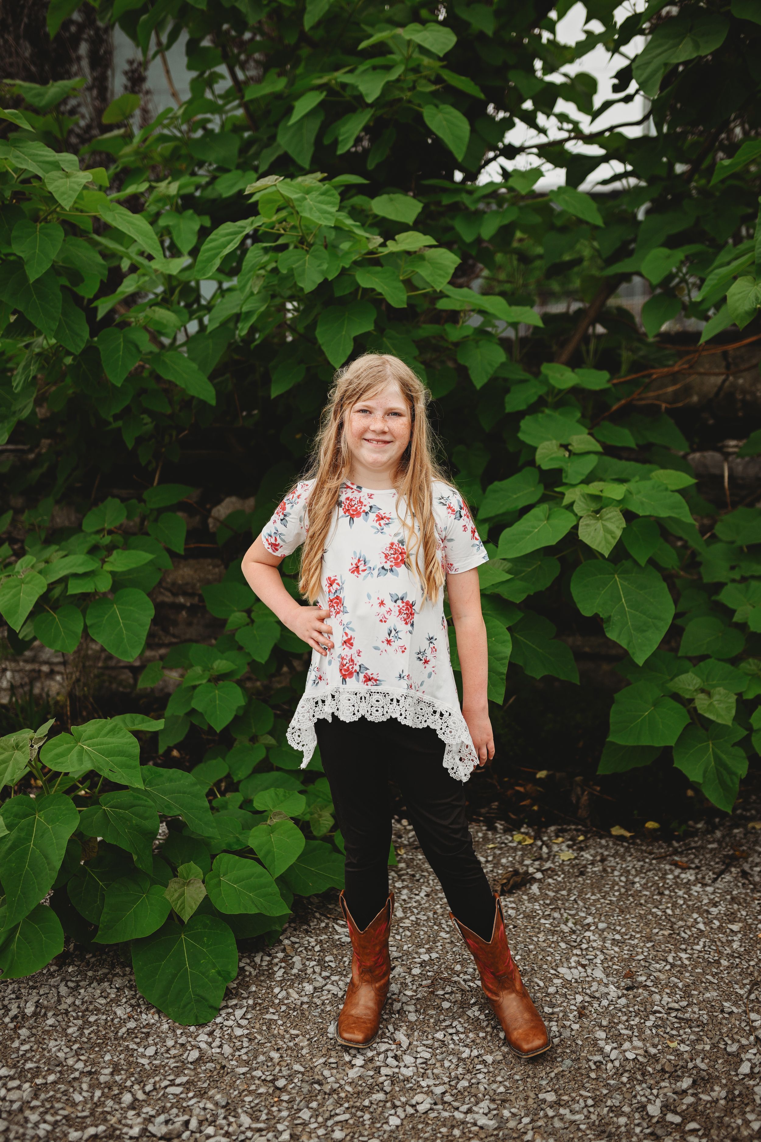 Child playing and posing on gravel path surrounded by lush green foliage.