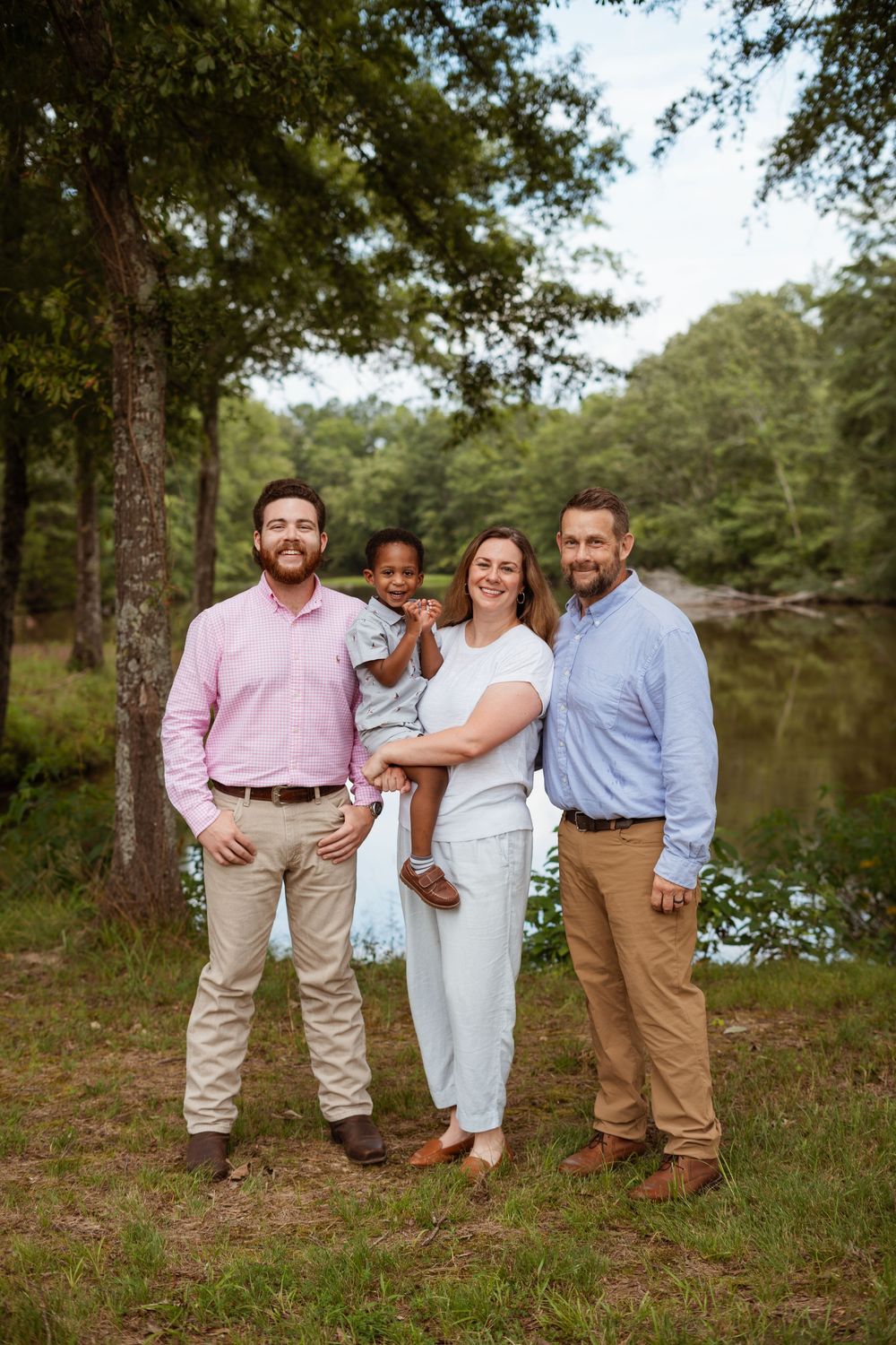 Group portrait of four people standing together by a tranquil pond in a wooded setting.