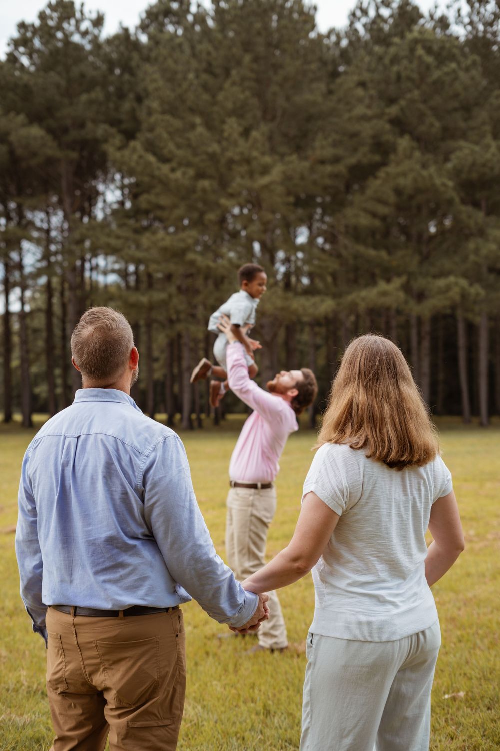 Family watching and holding hands as child is lifted high in the air in a grassy field.