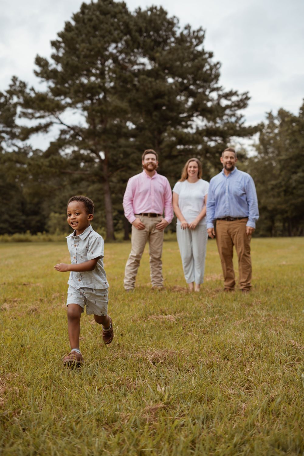 Child running joyfully in foreground with three adults standing in background near pine trees.