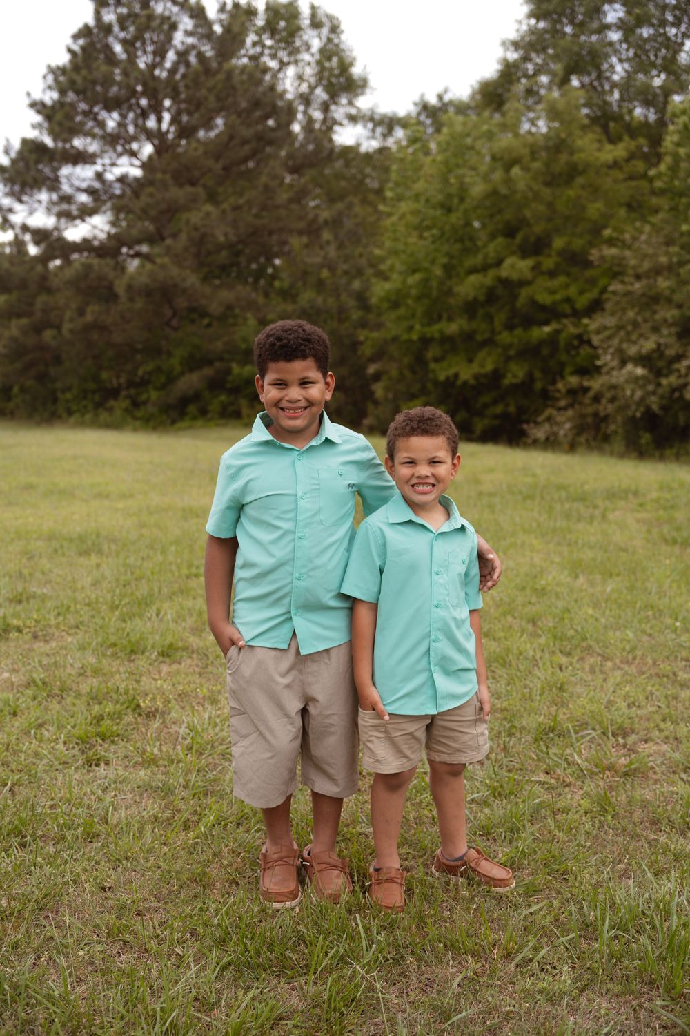 Two children in matching mint green shirts and khaki shorts pose together outdoors.