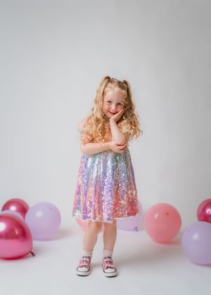 Young girl in rainbow sequin dress poses with pink balloons for birthday party photo.
