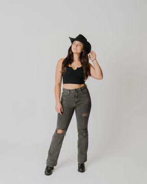 Young woman poses confidently in black cowboy hat, crop top, ripped gray jeans, and boots against neutral backdrop.