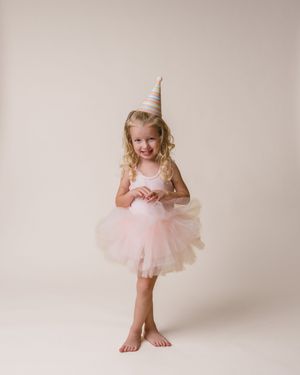 Adorable blonde girl twirls in pink tutu dress and striped party hat during birthday celebration portrait.