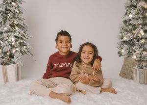 Two children smile together in Christmas sweaters beside decorated holiday trees in a festive studio portrait.