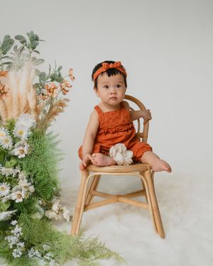 Adorable baby in rust orange romper sitting in a rattan chair surrounded by fresh flowers.