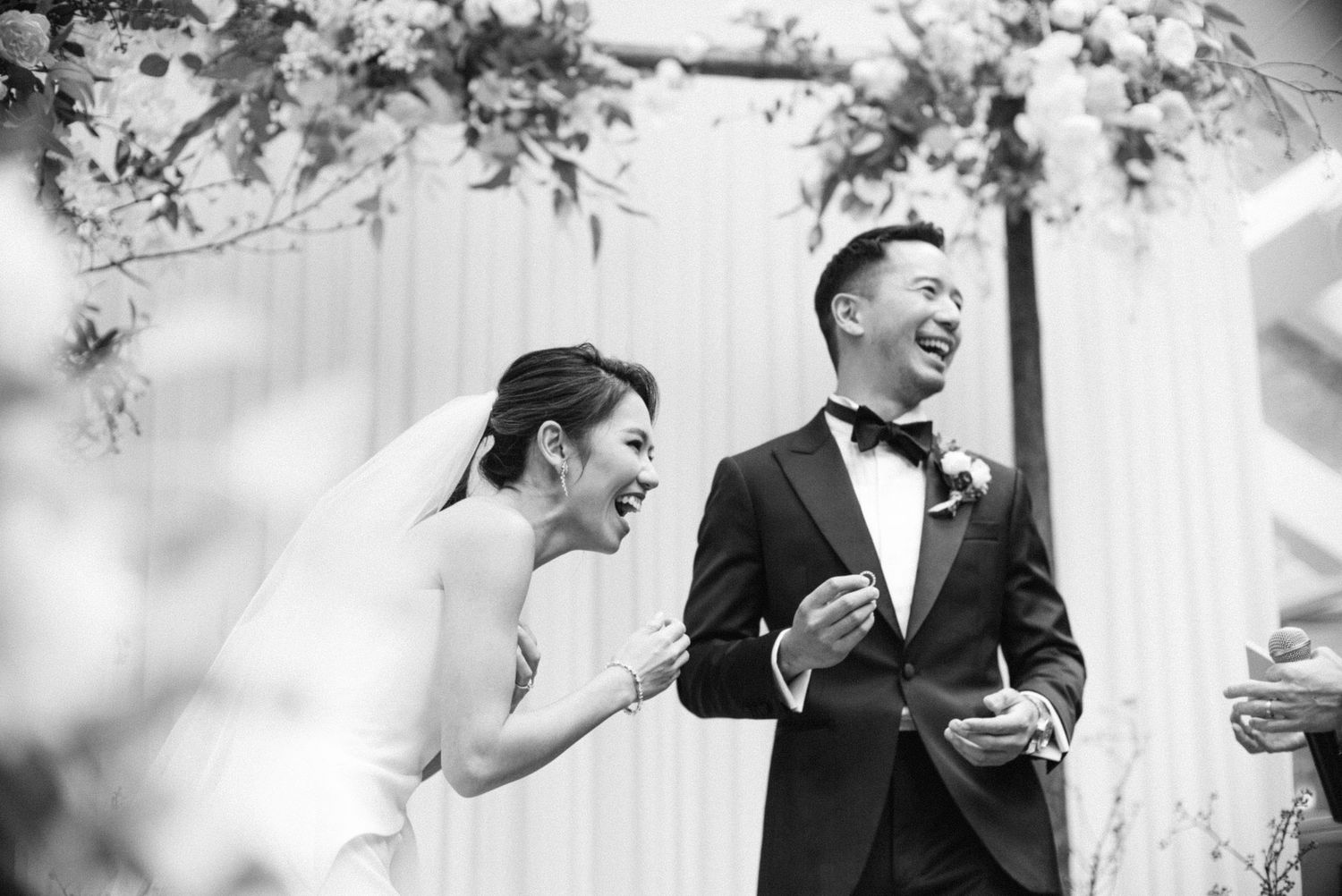 Black and white photo of a bride and groom laughing joyfully at their outdoor wedding ceremony.