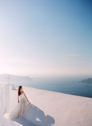 Bride in a flowing gown standing on white Santorini terrace overlooking the stunning Aegean Sea.