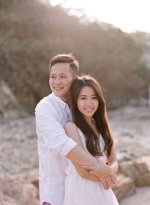 Smiling Asian couple embracing at golden hour on a rocky beach in casual white outfits.